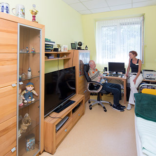 Ein Mann sitzt in einem Bürostuhl und spricht mit einer Frau, die neben ihm steht. Im Hintergrund sind ein Fernseher und ein Computer zu sehen. Der Raum ist hell und freundlich gestaltet, mit Regalen und Wohnmöbeln aus Holz.