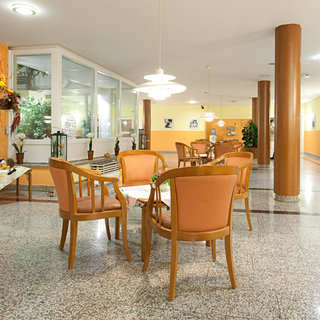 Eine helle, gemütliche Lounge mit mehreren Stühlen und Tischen. Die Einrichtung ist freundlich und einladend, mit Pflanzen und Dekorationen an den Wänden. Viel Tageslicht strömt durch die Fenster und schafft eine angenehme Atmosphäre.