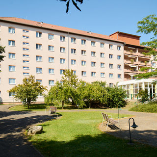 Ein mehrstöckiges Gebäude mit hellen Fassaden steht in einer grünen Umgebung. Vor dem Gebäude sind gepflegte Gärten mit Bäumen und Bänken, die eine einladende Atmosphäre schaffen. Der Himmel ist klar und blau.