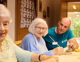 Eine Gruppe von älteren Menschen sitzt an einem Tisch und arbeitet mit Ton. Eine Frau mit Brille lächelt, während sie mit einem Werkzeug in ihrer Hand eine Figur formt. Ein Mann in einem blauen T-Shirt beobachtet sie freundlich. Im Hintergrund sind weitere Teilnehmer und Materialien sichtbar.