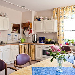 Die Küche ist hell und freundlich, mit weißen Schränken, einem Tisch mit blauer Tischdecke und einer Vase mit rosafarbenen Rosen. Ein Fenster mit gelben Vorhängen lässt viel Licht herein, und auf der Arbeitsplatte stehen Küchengeräte und Pflanzen.