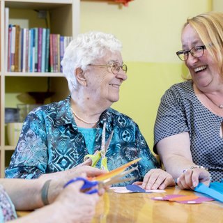 Drei Frauen, darunter eine ältere Dame mit weißen Haaren und Brille, sitzen an einem Tisch und lächeln. Sie sind mit Bastelmaterialien beschäftigt, darunter Scheren und farbige Papiere. Die Atmosphäre ist freundlich und humorvoll, was auf eine angenehme Zeit zusammen hindeutet.