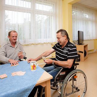 Zwei Männer sitzen an einem Tisch in einem hellen Raum. Der Mann im Rollstuhl zählt Geld, während der andere ihm aufmerksam zusieht. Auf dem Tisch liegt eine blaue Tischdecke und mehrere Karten. Beide Männer wirken entspannt und genießen ihre Zeit miteinander.