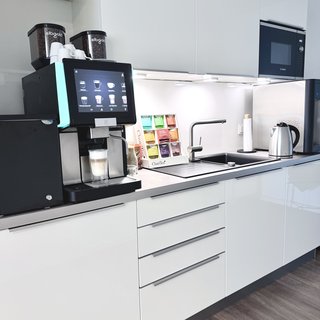 Eine moderne Küchenzeile mit einem Kaffeeautomaten, der ein Touchscreen-Display hat. Neben dem Automaten stehen verschiedene Kaffeepackungen in bunten Farben. Es gibt einen Wasserkocher und eine Spüle, alles in einem hellen, minimalistischen Design.