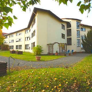 Ein modernes Gebäude mit mehreren Fenstern und einer hellen Fassade steht in einem gepflegten Garten. Um das Gebäude herum sind grüne Rasenflächen, kleine Bäume und Sitzmöglichkeiten angeordnet. Die Atmosphäre wirkt einladend und ruhig.