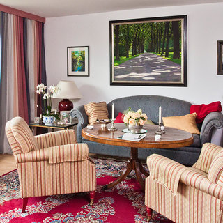 Ein gemütlicher Wohnraum mit zwei gestreiften Sesseln und einem runden Tisch in der Mitte. Auf dem Tisch stehen Blumen und einige dekorative Gegenstände. An den Wänden hängen Bilder, und ein großes Fenster lässt natürliches Licht herein. Ein Teppich vervollständigt das harmonische Ambiente.