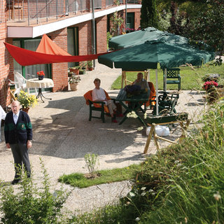 Eine Person in einer weißen Jacke steht neben einer älteren Dame in einem begrünten Innenhof mit Sitzgelegenheiten. Im Hintergrund sitzen drei weitere Personen unter einem großen Sonnenschirm. Es ist sonnig und die Atmosphäre wirkt einladend und freundlich.