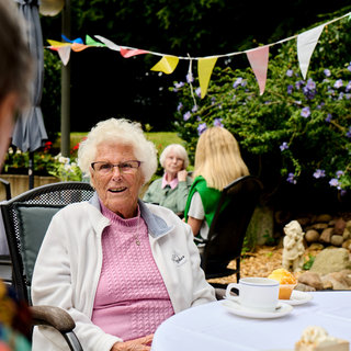 Eine ältere Frau mit weißem Haar und Brille sitzt an einem Tisch im Freien, lächelt und trägt eine rosa Strickjacke. Vor ihr stehen eine Tasse Kaffee und ein Muffin. Im Hintergrund sind weitere Personen und dekorative Wimpel sichtbar.