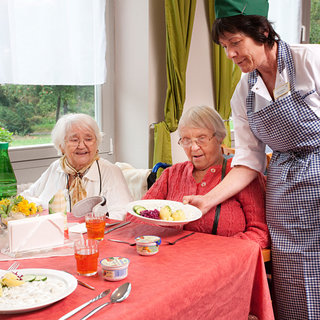 Drei ältere Frauen sitzen an einem Tisch mit gedeckter Tafel. Eine Frau in einer Kochschürze serviert eine Mahlzeit. Auf dem Tisch stehen verschiedene Gerichte und Getränke. Die Atmosphäre wirkt freundlich und einladend.