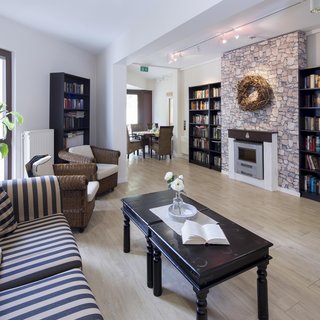 Ein gemütlicher Raum mit einem modernen Bücherregal, das von Boden bis zur Decke reicht. In der Mitte steht ein schwarzer Tisch mit einem aufgeschlagenen Buch und einer Vase mit einer Blume. Ein komfortables Sofa und ein Sessel laden zum Lesen ein.