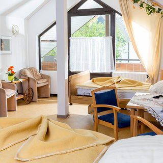Ein helles Zimmer mit zwei Pflegebetten, einem Tisch mit Stühlen und einer gemütlichen Sitzecke. Fenster mit Vorhängen lassen viel Tageslicht herein, und es sind einige Pflanzen und persönliche Gegenstände im Raum. Eine warme und einladende Atmosphäre herrscht vor.