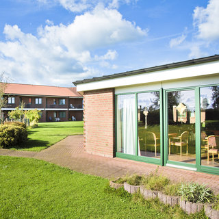 Ein modernes Gebäude mit großen Fenstern und einer Terrasse blickt auf einen gepflegten Garten. Im Hintergrund sind weitere Wohngebäude zu sehen, umgeben von Grünflächen und Bäumen. Der Himmel ist klar mit wenigen Wolken.