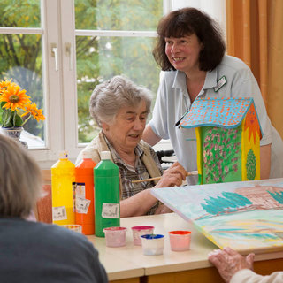 Eine ältere Frau malt an einem Tisch, während eine Betreuerin sie anlächelt und unterstützt. Auf dem Tisch stehen Becher mit bunten Farben und eine bereits bemalte, farbenfrohe Vogelhausfigur. Im Hintergrund sind frische Blumen zu sehen.