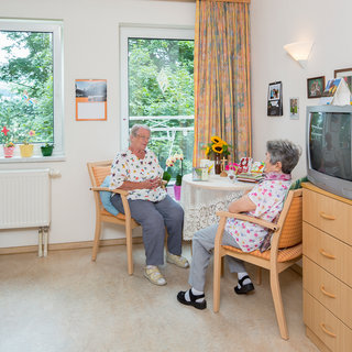 Zwei ältere Frauen sitzen an einem Tisch in einem hellen Zimmer. Sie unterhalten sich freundlich, umgeben von Pflanzen und persönlichen Gegenständen. Sonnenschein fällt durch das Fenster und schafft eine einladende Atmosphäre.