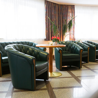 In einem hellen Raum stehen vier grüne, gepolsterte Sessel um einen runden Tisch. Eine Pflanze und einige Blumen sind ebenfalls im Hintergrund sichtbar. Die Fenster lassen viel Tageslicht herein, was eine einladende Atmosphäre schafft.