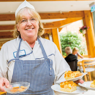 Eine lächelnde Servicekraft in Kochuniform trägt mehrere Teller mit Kuchen und Gebäck. Hintergrund: eine einladende Teestube mit Gästen, die entspannt sitzen. Das Bild vermittelt eine freundliche Atmosphäre des Genießens und der Gastfreundschaft.