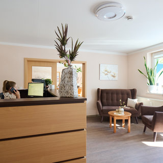 Eine helle Empfangshalle mit einem Holzschreibtisch, an dem eine Mitarbeiterin telefoniert. Links steht ein Computer. In der Mitte befindet sich ein kleiner Tisch mit einer Blumenvase. Rechts sind zwei bequeme Sessel und Pflanzen vor einem Fenster.