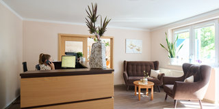Eine helle Empfangshalle mit einem Holzschreibtisch, an dem eine Mitarbeiterin telefoniert. Links steht ein Computer. In der Mitte befindet sich ein kleiner Tisch mit einer Blumenvase. Rechts sind zwei bequeme Sessel und Pflanzen vor einem Fenster.