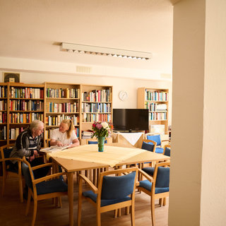 Eine ältere Dame und ein älterer Herr sitzen an einem Tisch in einer Bibliothek mit Regalen voller Bücher. Auf dem Tisch steht eine Vase mit Blumen. Im Hintergrund ist ein Fernseher und ein Fenster mit Tageslicht zu sehen.