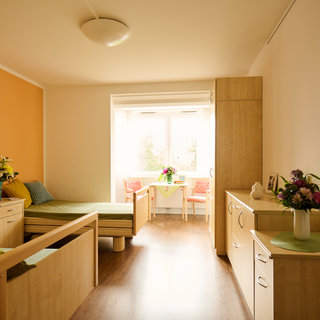 Ein helles Zimmer mit einem orangefarbenen Akzent an der Wand. Es gibt zwei Betten, eine Schrankeinheit, einen Tisch mit Stühlen und Blumenarrangements in Vasen. Ein Fenster lässt Tageslicht herein.