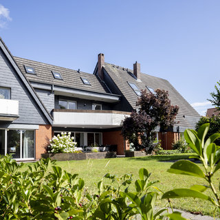 Ein modernes Mehrfamilienhaus mit einer Kombination aus roten Ziegeln und dunklen Schindeln. Das Gebäude hat ein begrüntes Grundstück mit Bäumen und Sträuchern. Im Vordergrund sind Pflanzen sichtbar, die zur Umgebung des Hauses gehören. Der Himmel ist klar und blau.