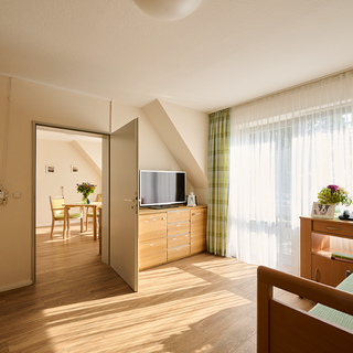 Ein modernes Zimmer mit Holzfußboden, einem Fenster mit Vorhängen, einem TV auf einem Schrank und einem Bett. Durch eine offene Tür ist ein Essbereich mit Tisch und Stühlen sichtbar. Pflanzen bringen eine einladende Atmosphäre.
