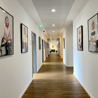 Ein heller Gang mit Holzfußboden, der von Rahmungen mit Fotografien älterer Menschen gesäumt ist. Die Bilder zeigen verschiedene Personen, die freundlich in die Kamera blicken, und verleihen dem Raum eine einladende Atmosphäre.