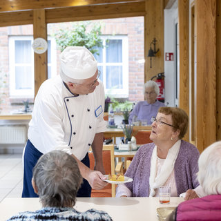 Ein Kellner in Kochuniform reicht einer älteren Dame am Tisch einen Teller mit Essen. Im Hintergrund sind weitere Senioren zu sehen. Die Atmosphäre wirkt freundlich und einladend.