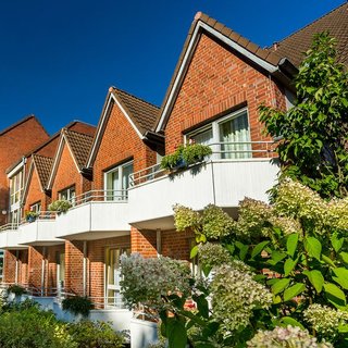 Eine Ansicht von zwei dreigeschossigen, roten Ziegelhäusern mit spitzen Dächern. Die Balkone sind mit Blumen dekoriert, und im Vordergrund blühen Sträucher. Der Himmel ist klar und blau, was eine freundliche und einladende Atmosphäre vermittelt.