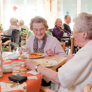 Zwei ältere Frauen sitzen an einem Tisch in einem Speisesaal. Eine Frau lächelt und hält einen Teller mit Essen, während die andere ihr aufmerksam zuschaut. Im Hintergrund sind weitere Personen und Essensbereiche zu sehen, die eine gesellige Atmosphäre schaffen.
