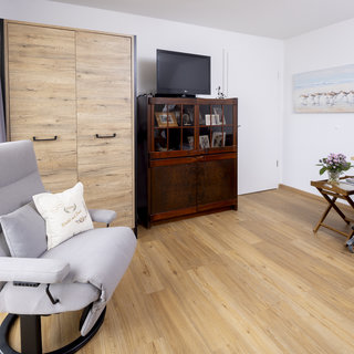 Ein helles Zimmer mit Holzboden. Links steht ein grauer Relaxsessel mit einem dekorativen Kissen. Gegenüber gibt es einen dunklen Schrank mit Glastüren, in dem Bilder zu sehen sind. Rechts befindet sich ein Pflegebett mit gestreifter Bettwäsche und einem kleinen Tisch, auf dem ein Blumenstrauß steht.