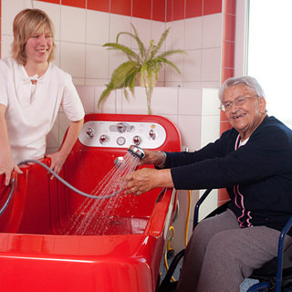 Eine Pflegerin steht neben einer roten Badewanne und hilft einer älteren Frau im Rollstuhl, während Wasser aus einem Duschkopf spritzt. Die Frau lächelt zufrieden und genießt die Unterstützung beim Baden. Im Hintergrund ist eine Pflanze und eine Fensterbank sichtbar.