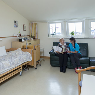 Eine ältere Frau sitzt auf einem Sofa und liest ein Buch, während eine jüngere Frau, die neben ihr sitzt, aufmerksam zuhört. Der Raum ist hell und freundlich, mit einem Bett, einem Tisch und Pflanzen in den Fenstern.