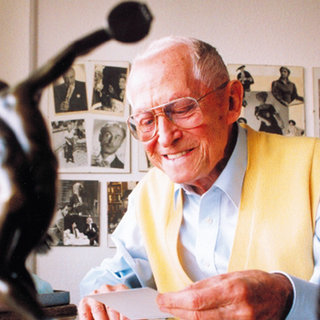 Ein älterer Mann mit Brille und einem gelben Pullover sitzt an einem Tisch und lächelt. Er hält eine Karte in der Hand. Im Hintergrund sind schwarz-weiße Fotos an der Wand zu sehen. Eine kleine Statue ist im Vordergrund.