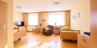 Ein heller und freundlicher Raum mit zwei Betten, einem Tisch und Stühlen. Auf der linken Seite steht ein Fernseher. Ein Sessel und eine Couch sind ebenfalls sichtbar. Große Fenster mit Vorhängen lassen viel Licht herein. Der Boden ist lichtbraun und der Raum wirkt einladend.