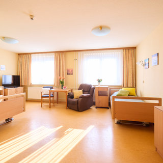 Ein heller und freundlicher Raum mit zwei Betten, einem Tisch und Stühlen. Auf der linken Seite steht ein Fernseher. Ein Sessel und eine Couch sind ebenfalls sichtbar. Große Fenster mit Vorhängen lassen viel Licht herein. Der Boden ist lichtbraun und der Raum wirkt einladend.