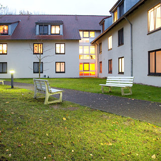 Eine ruhige Gartenanlage vor einem Gebäude. Es gibt mehrere Bänke, einen beleuchteten Lampenpfosten und einen gepflasterten Weg. Die Fenster des Gebäudes strahlen warmes Licht aus, während die Dämmerung einsetzt. Die Atmosphäre wirkt einladend und friedlich.