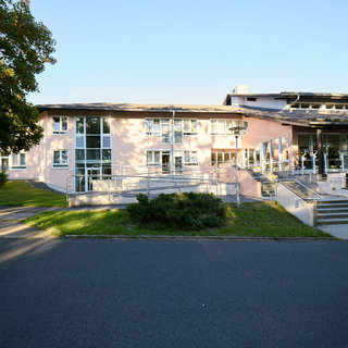 Ein modernes, eingeschossiges Gebäude in hellen Farben mit großen Fenstern und einem darüberliegenden Vordach. Es gibt einen Zugang mit Stufen und eine Rampe für barrierefreies Betreten. Im Vordergrund ist ein begrüntes Areal mit Bäumen und einem gepflasterten Weg zu sehen.