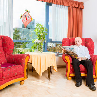 Ein älterer Mann sitzt in einem roten Sessel und liest eine Zeitung. Neben ihm steht ein runder Tisch mit einer Tischdecke. Im Hintergrund sind zwei große Fenster mit Vorhängen und Pflanzen zu sehen. Das Licht ist hell und freundlich.