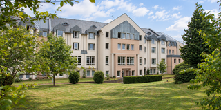 Das Bild zeigt ein modernes Wohngebäude mit mehreren Fenstern und einem begrünten Gartenbereich. Umgeben von Bäumen und Sträuchern, erstreckt sich eine gepflegte Rasenfläche vor dem Gebäude, unter einem blauen Himmel mit einigen Wolken.