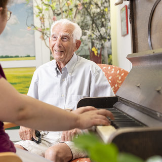 Eine ältere Frau spielt auf einem Klavier, während ein lächelnder, älterer Mann ihr aufmerksam zuhört. Beide sitzen in einem hellen Raum mit bunten Bildern an den Wänden. Die Atmosphäre ist freundlich und entspannt.