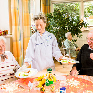 Eine Mitarbeiterin serviert zwei Senioren in einem Essraum Mahlzeiten. Der Tisch ist gedeckt, und hinter den Senioren sind weitere Bewohner zu sehen. Die Atmosphäre wirkt freundlich und einladend.