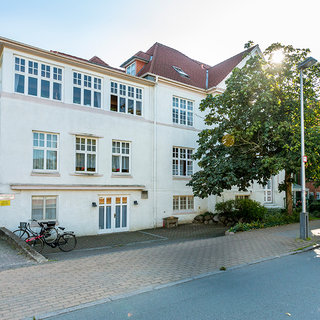 Das Bild zeigt ein mehrstöckiges, weißes Wohngebäude mit großen Fenstern. Vor der Fassade steht ein Baum, und daneben befindet sich ein Fahrrad. Die Aufnahme ist bei Tageslicht gemacht und vermittelt eine ruhige, städtische Atmosphäre.