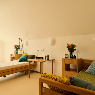 Ein heller und freundlicher Raum mit zwei Betten. Neben den Betten stehen Nachttische mit Dekoration, darunter Blumen und einige persönliche Gegenstände. Ein Fenster lässt Tageslicht herein und verleiht dem Raum eine einladende Atmosphäre.