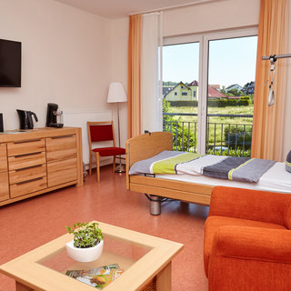 Das Bild zeigt ein helles, freundliches Zimmer mit einem Pflegebett, einem TV, einem bequemen Sessel und einem Tisch mit einer Pflanze. Große Fenster bieten Ausblick auf eine grüne Umgebung. Der Raum vermittelt einladende, wohnliche Atmosphäre.
