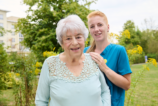 Eine ältere Frau mit grauen Haaren lächelt in die Kamera. Hinter ihr steht eine junge, blonde Frau in blauer Arbeitskleidung und legt ihre Hände auf die Schultern der Seniorin. Sie befinden sich in einem Garten mit Pflanzen und Bäumen im Hintergrund.