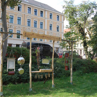 Ein Holzspielgerät im Freien, das verschiedene Musikinstrumente wie Trommeln und Metallklänge enthält. Es steht umgeben von Rasen und Bäumen, mit einem Gebäude im Hintergrund. Das Gerät lädt Kinder zum Musizieren und kreativen Spielen ein.