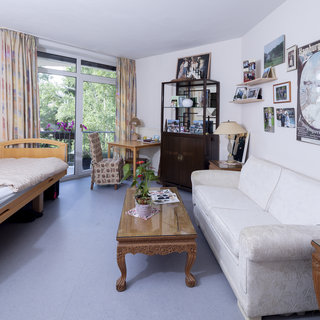 Das Zimmer ist hell und freundlich eingerichtet, mit einem Bett auf der linken Seite, einem weißen Sofa und einem Tisch am Fenster. An den Wänden hängen Bilder und Fotografien. Ein Pflanztopf steht auf dem Tisch, und der Blick geht nach draußen auf die Natur.