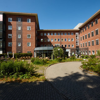 Ein mehrstöckiges, modernes Gebäude aus roten Ziegelsteinen mit großen Fenstern umgeben von einem gepflegten Garten. Im Vordergrund ist ein gewundener Weg, der durch Blumenbeete führt. Die Szenerie ist bei sonnigem Wetter aufgenommen.