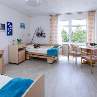 Ein helles Zimmer mit zwei Betten, einem Tisch und Stühlen. An der Wand hängen Bilder. Vor dem Fenster sind Pflanzen auf einem Tisch. Der Raum vermittelt eine freundliche und einladende Atmosphäre.